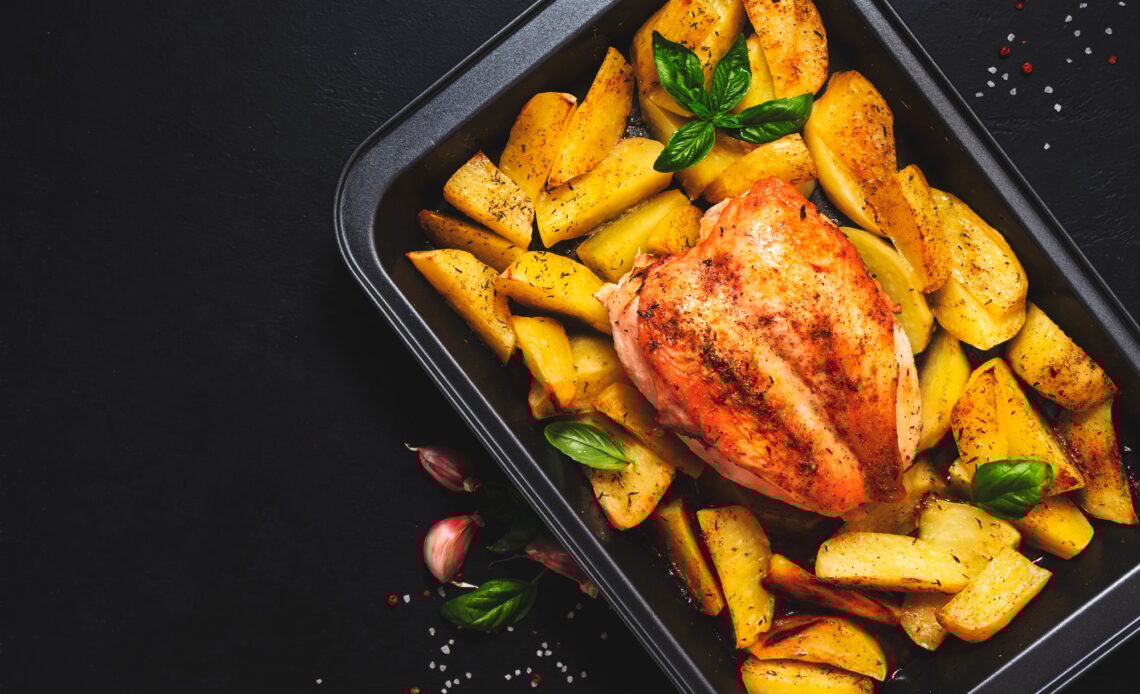 Baked chicken breast with golden potatoes in baking dish on dark background, top view