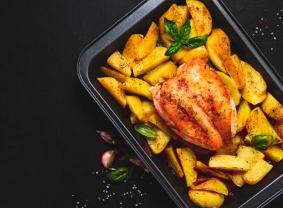 Baked chicken breast with golden potatoes in baking dish on dark background, top view