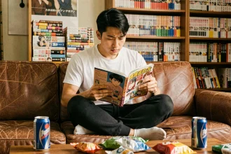 Jeune homme lisant un manga One Punch Man dans un appartement cosy entouré de volumes empilés sur des étagères